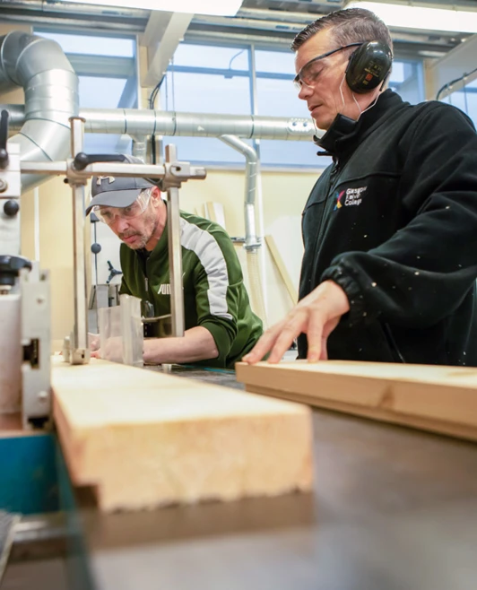 Two joinery lecturers in a woodworking workshop operating machinery, carefully guiding timber through cutting equipment, wearing safety gear. Two joinery lecturers in a woodworking workshop operating machinery, carefully guiding timber through cutting equipment, wearing safety gear.